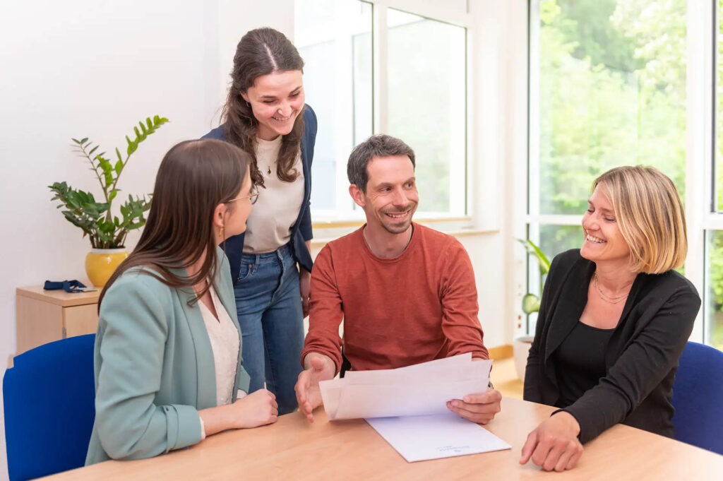 Vier Personen im Gespraech, laechelnd, mit Dokumenten am Tisch.